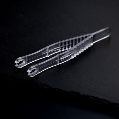 Zwei transparente, sterile Runde Piercingzangen liegen überkreuzt auf einem dunklen Hintergrund.